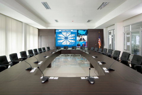 An executive conference room with a long table, individual microphones, and a large display screen in the front of the room. 