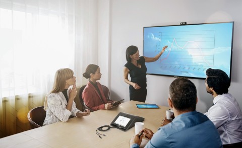 A team is holding a meeting in a conference room with a presenter motioning to a chart on the display in the front of the room.